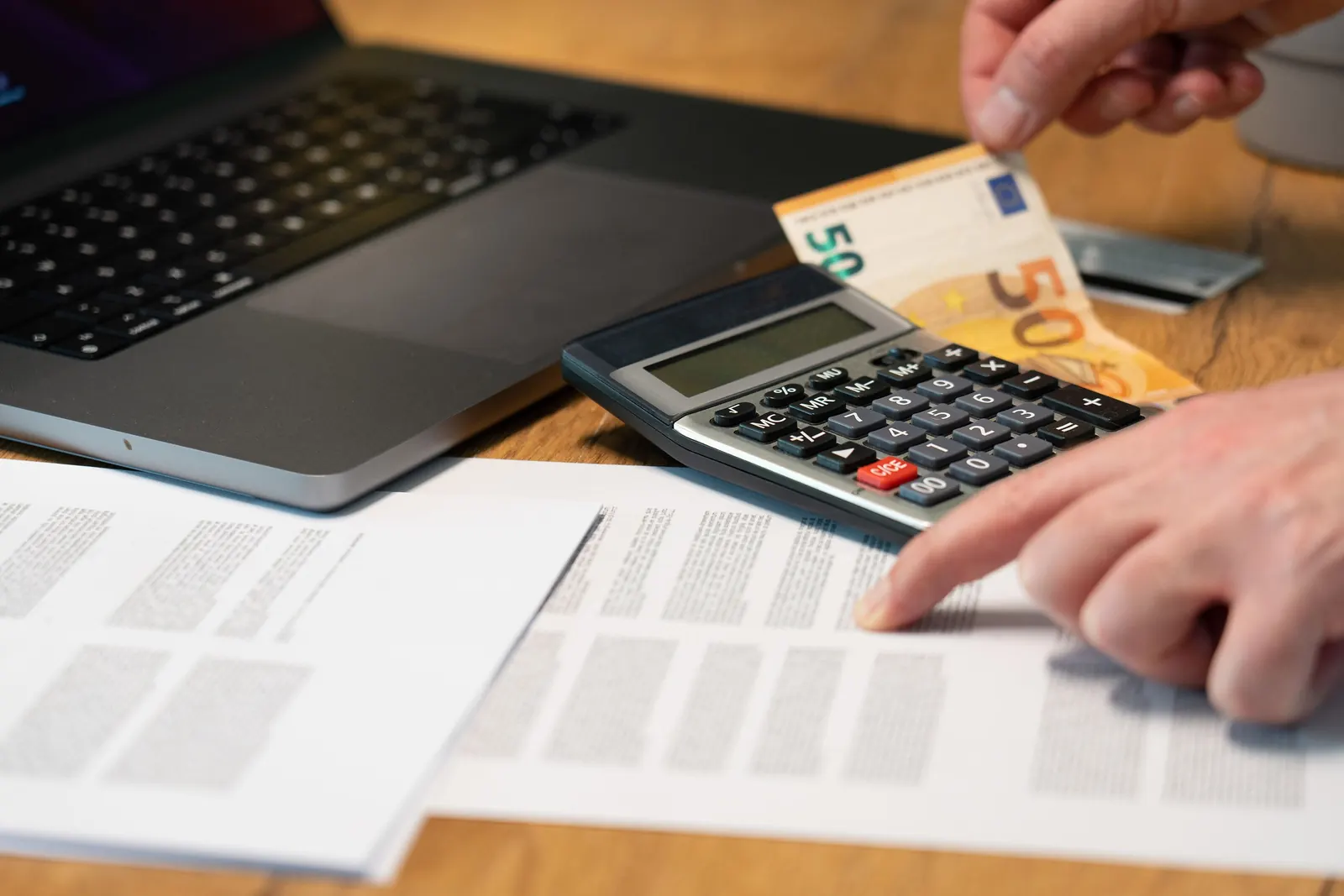 Mani che usano una calcolatrice su fogli di conti, con banconote da 50 euro e un portatile sulla scrivania