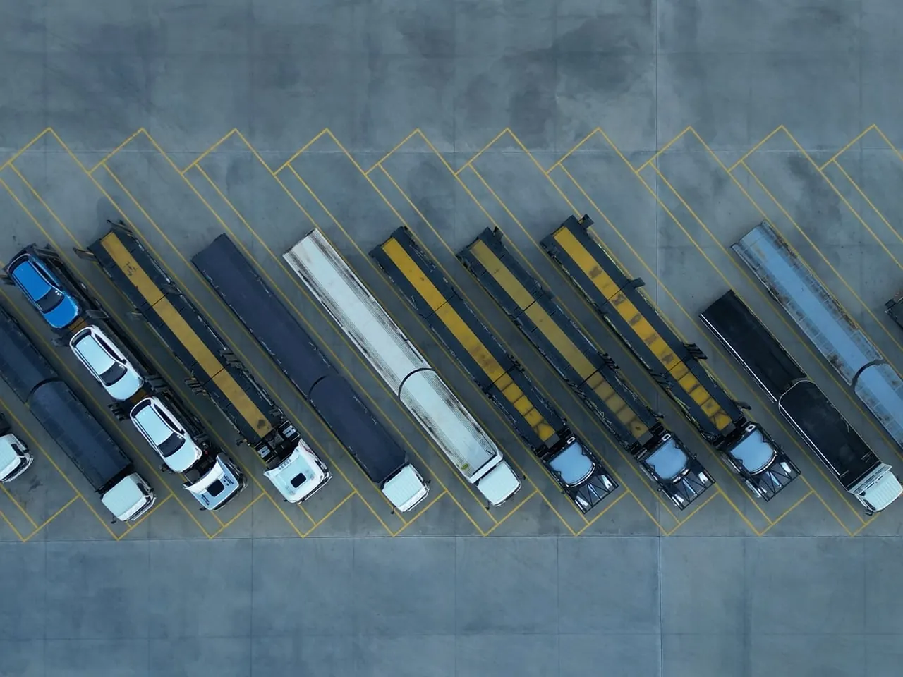 Vista dall'alto di una fila di camion e bisarche parcheggiati in diagonale su un piazzale con strisce gialle