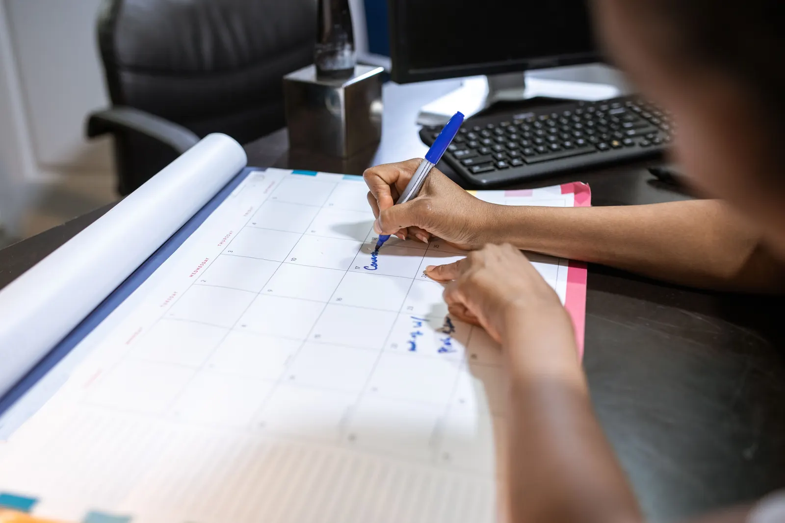 Mano che segna una scadenza con un pennarello blu su un grande calendario da scrivania, con tastiera e monitor sullo sfondo